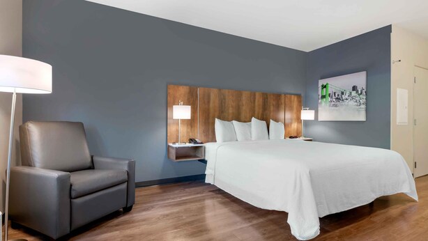 A hotel room with a large bed, a grey armchair, a bedside table with a lamp, and a wall-mounted shelf. - Extended Stay America Suites Charlotte Pineville Park Rd (Pineville)