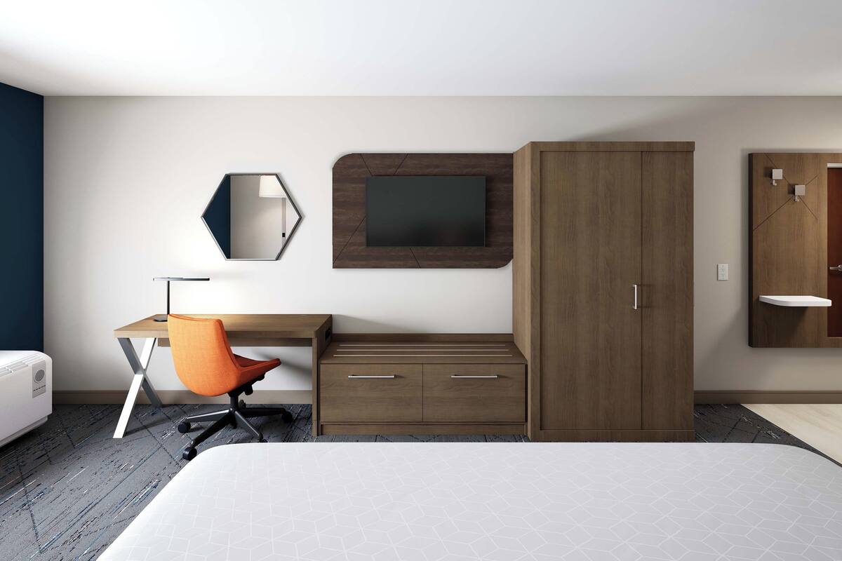 A modern hotel room with a bed, a desk with a chair, a wardrobe, a TV, and a mirror.