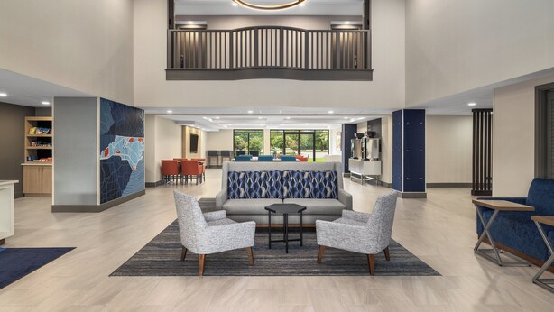 A spacious lobby with a high ceiling, a central seating area with a sofa and chairs, a kitchen area in the background, and a view of greenery outside. - Holiday Inn Express Fayetteville – Fort Bragg by IHG (Fayetteville)