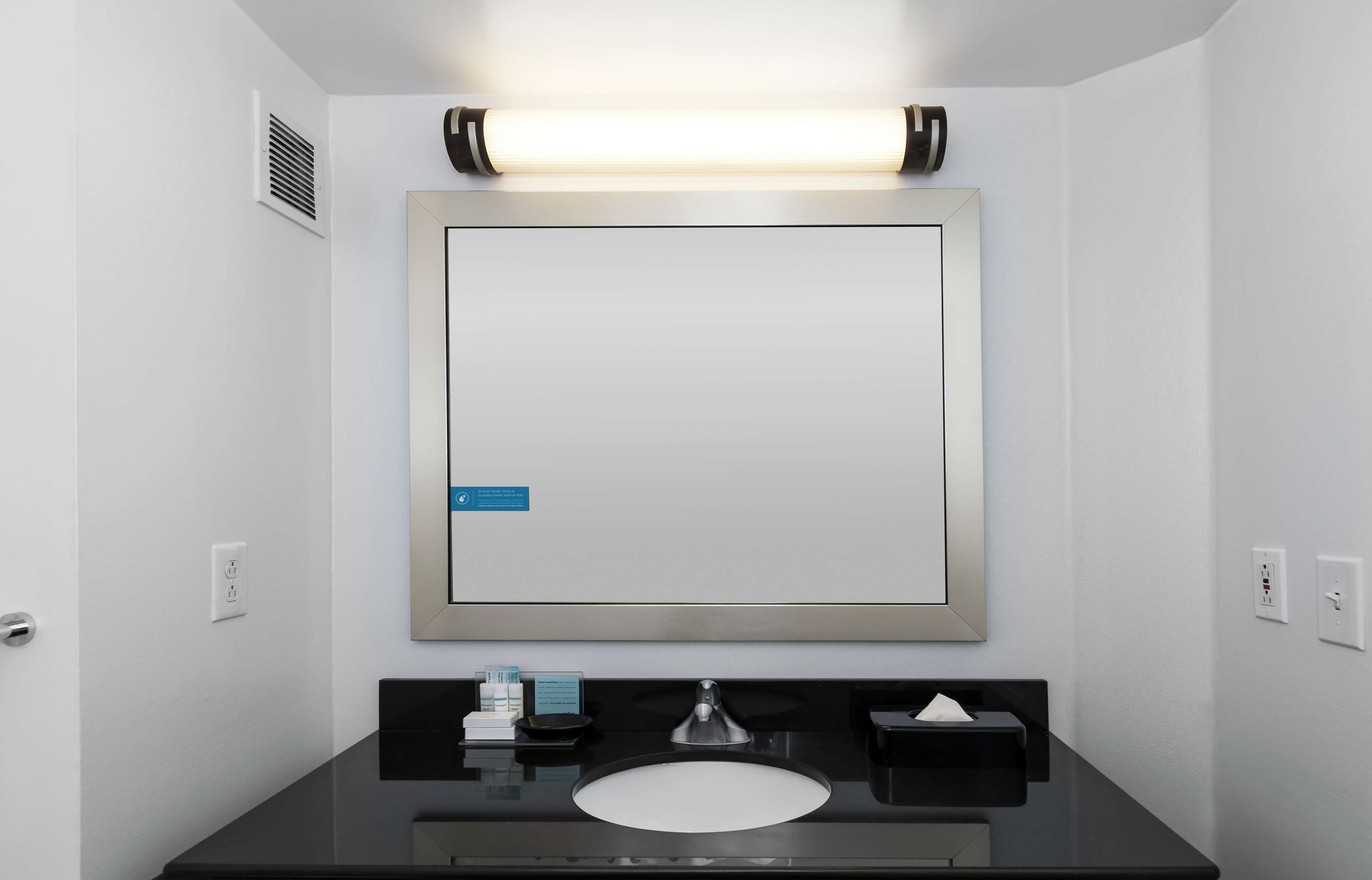 Un bagno moderno con uno specchio grande, un lavabo e un mobile contenitore con specchio. Sopra lo specchio c'è un lampadario e una bocchetta di areazione sulla parete.