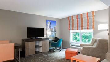 A hotel room with a desk, a television, a chair, a sofa, and a window with blinds.