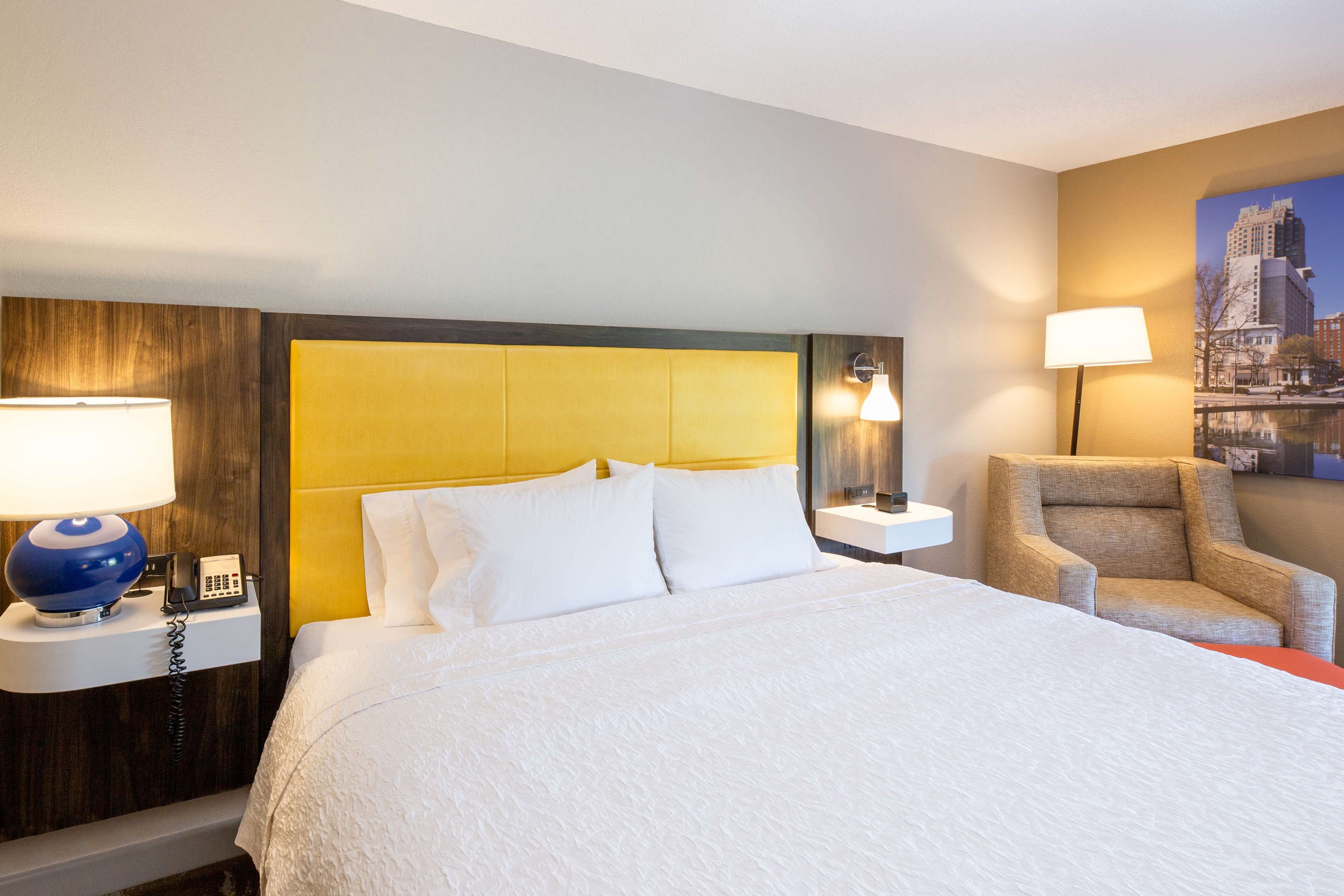 A hotel room with a large bed, a wooden headboard, a nightstand with a lamp, and a chair.