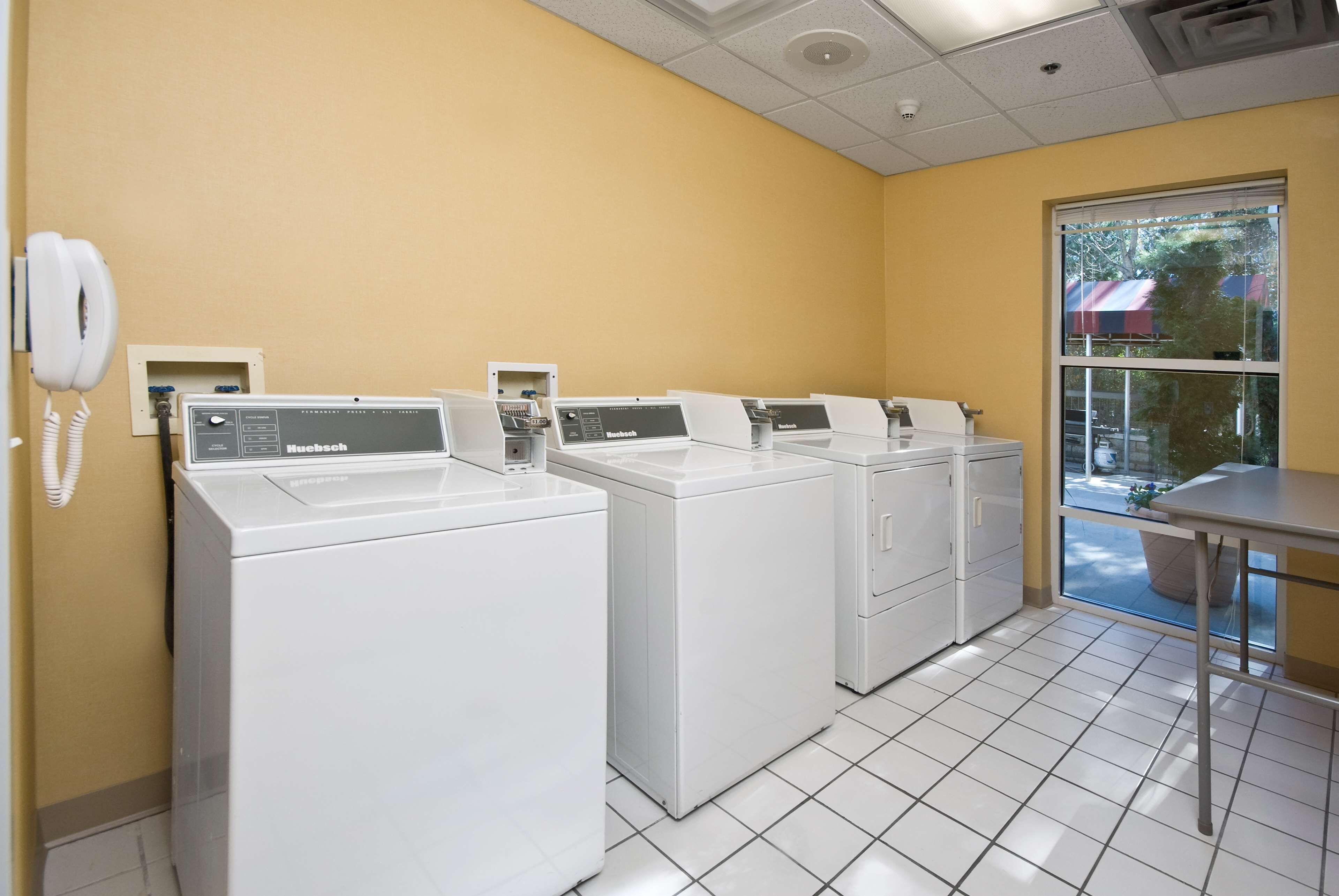 A laundry room with four white front-loading washers and dryers, a phone, and a window with a view of the outside.