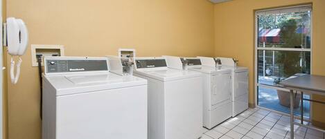 A laundry room with four white front-loading washers and dryers, a phone, and a window with a view of the outside.