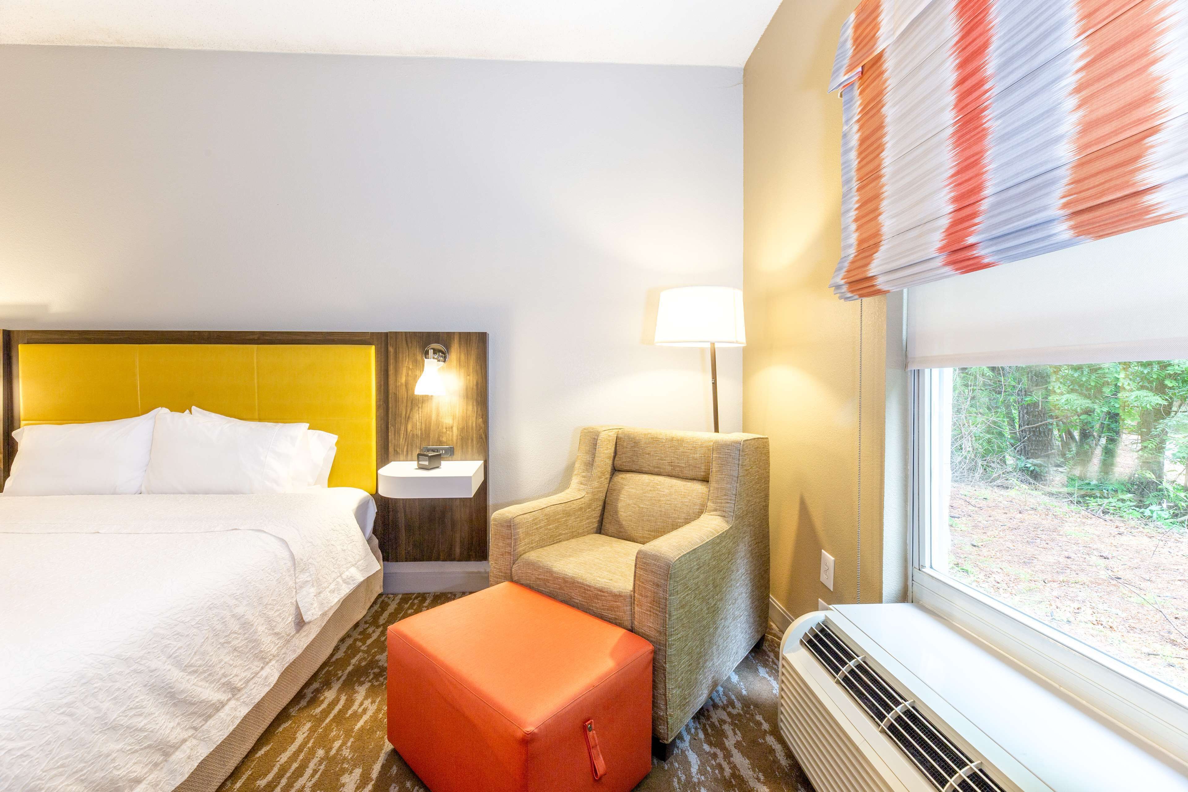 Une chambre d’hôtel comprenant un lit, une chaise, un pouf, une table de chevet, une lampe et une fenêtre donnant sur des arbres.