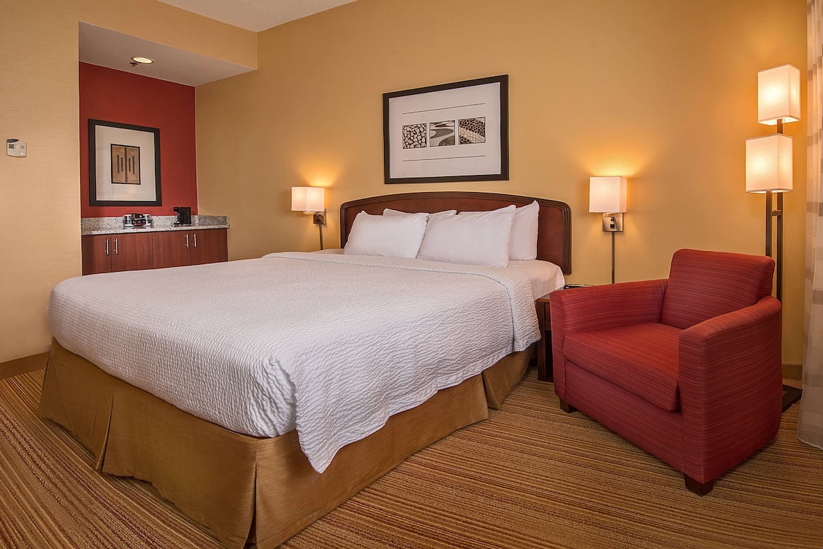 A hotel room with a large bed, a red armchair, a small kitchenette, and two wall-mounted lamps.