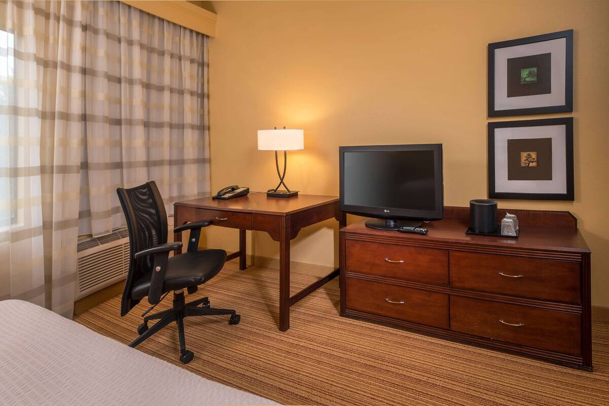 A hotel room with a bed, a desk with a computer, a chair, a lamp, a TV, and a dresser.