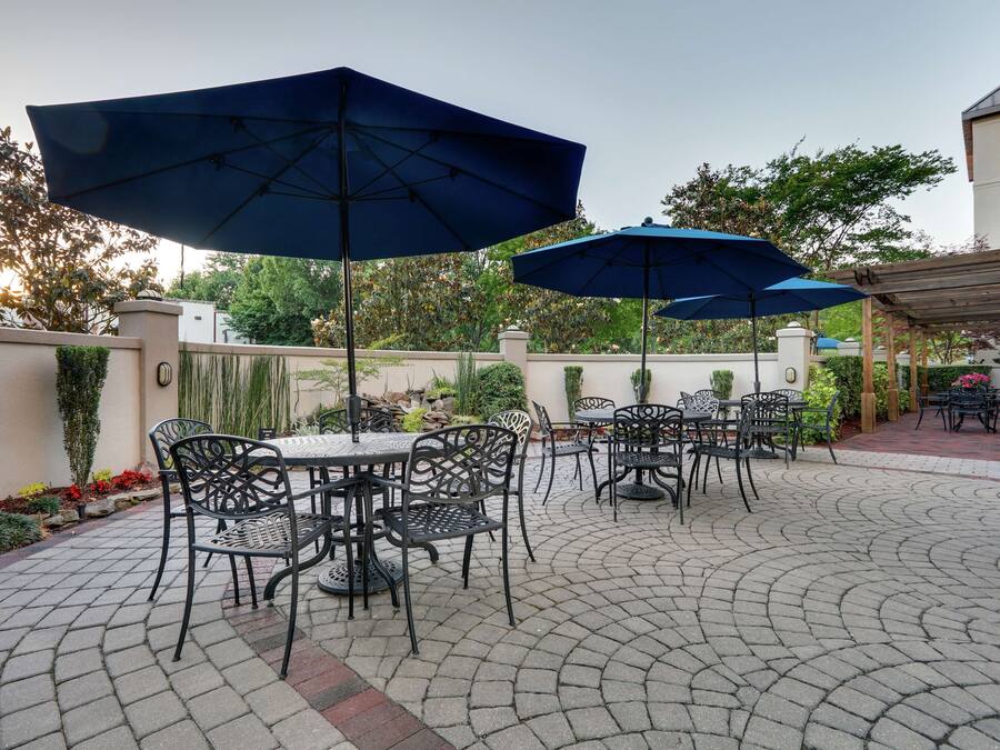 An outdoor patio with tables and chairs, covered by blue umbrellas, and surrounded by greenery.