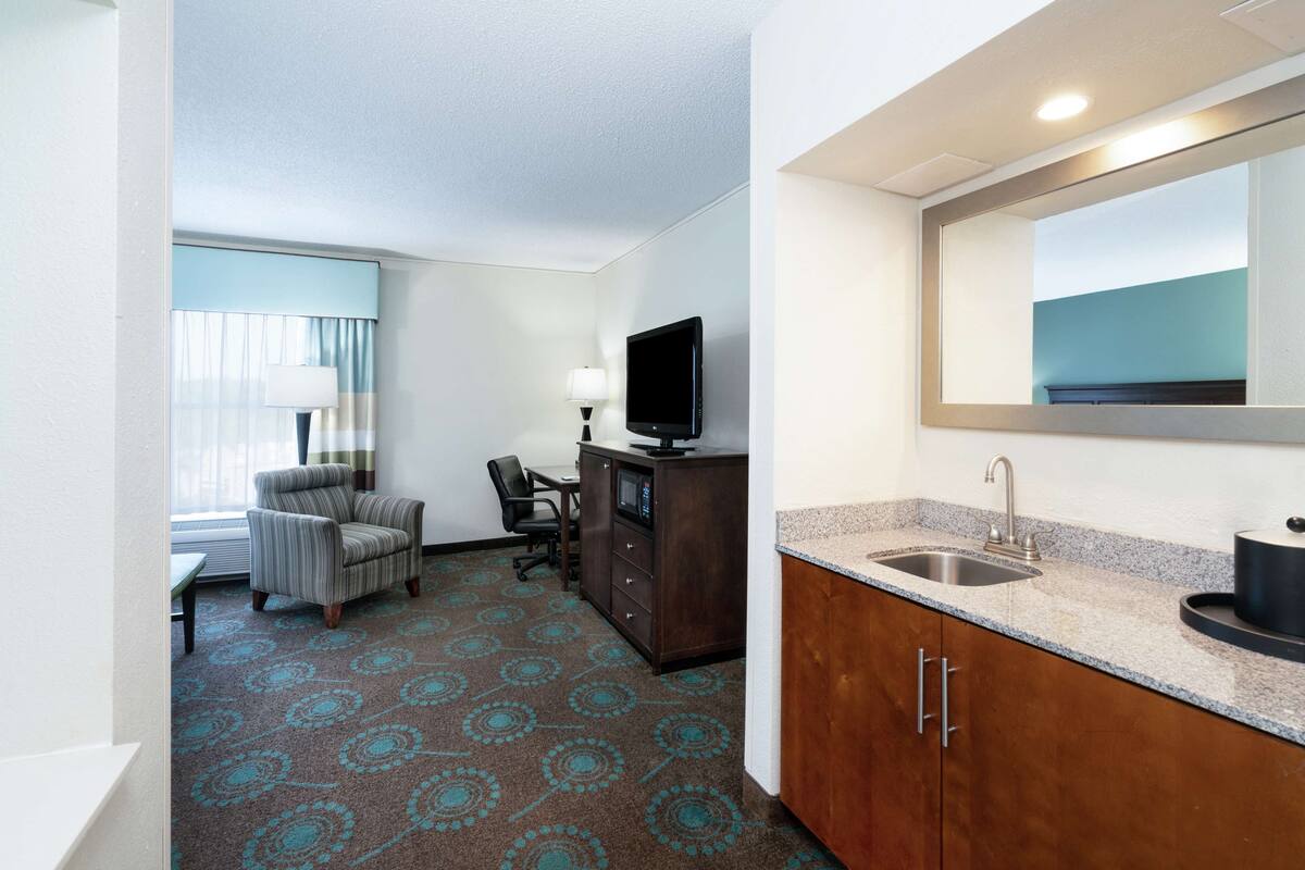 A hotel room with a kitchenette, a television, a desk, a chair, and a patterned carpet.