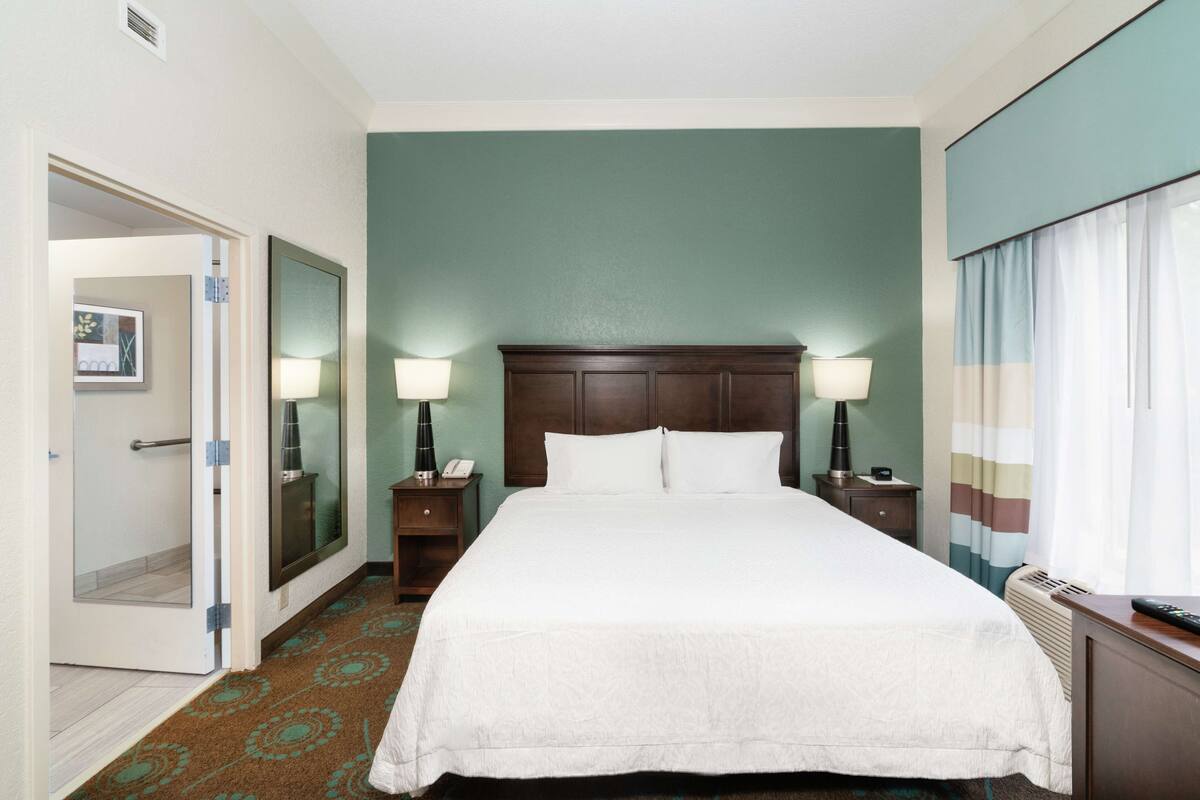A hotel room with a large bed, two bedside lamps, a nightstand, a mirror, and a view of the outdoors through a window with curtains.