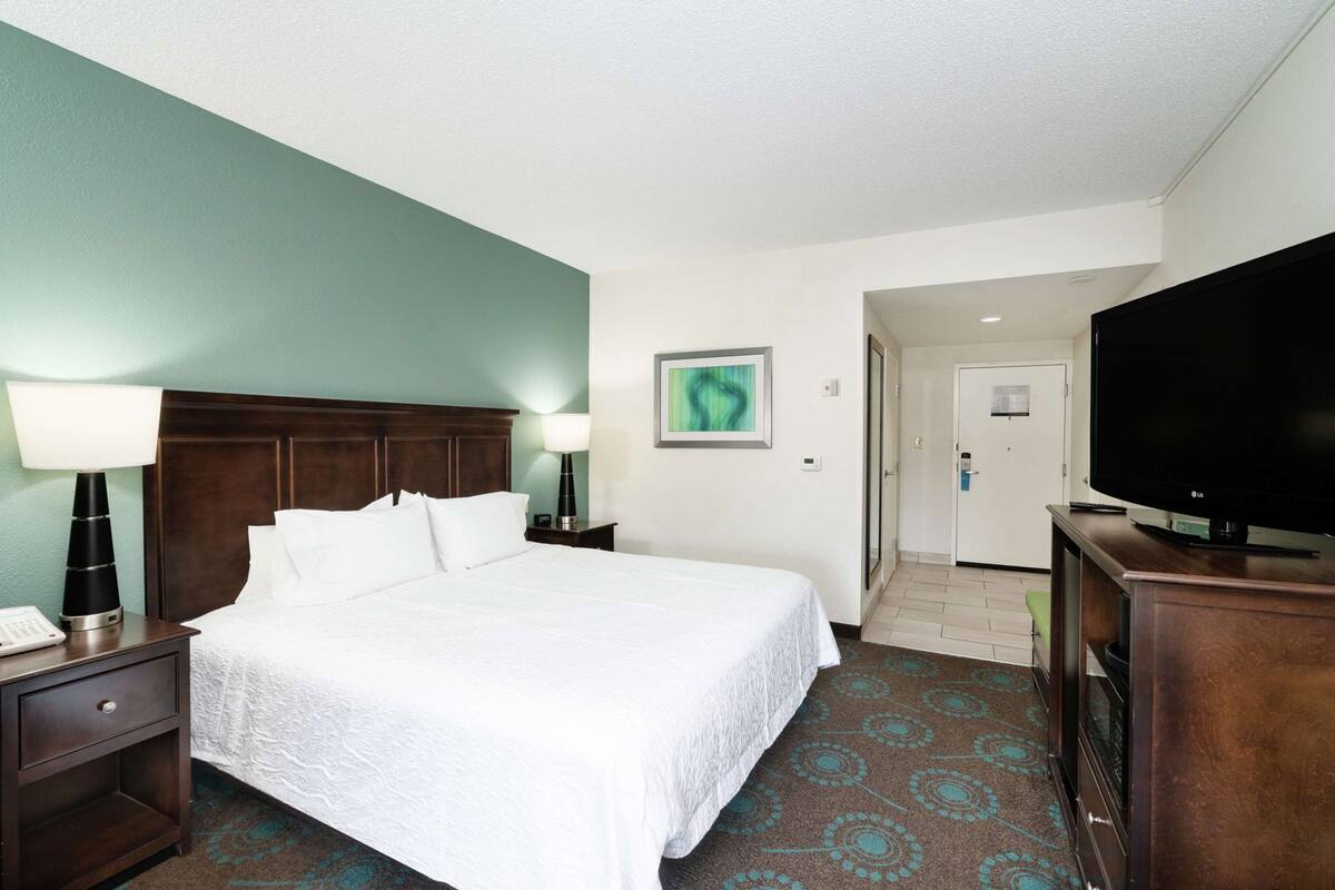 A hotel room with a bed, a TV, a desk with a lamp, a painting on the wall, and a door leading to another room.