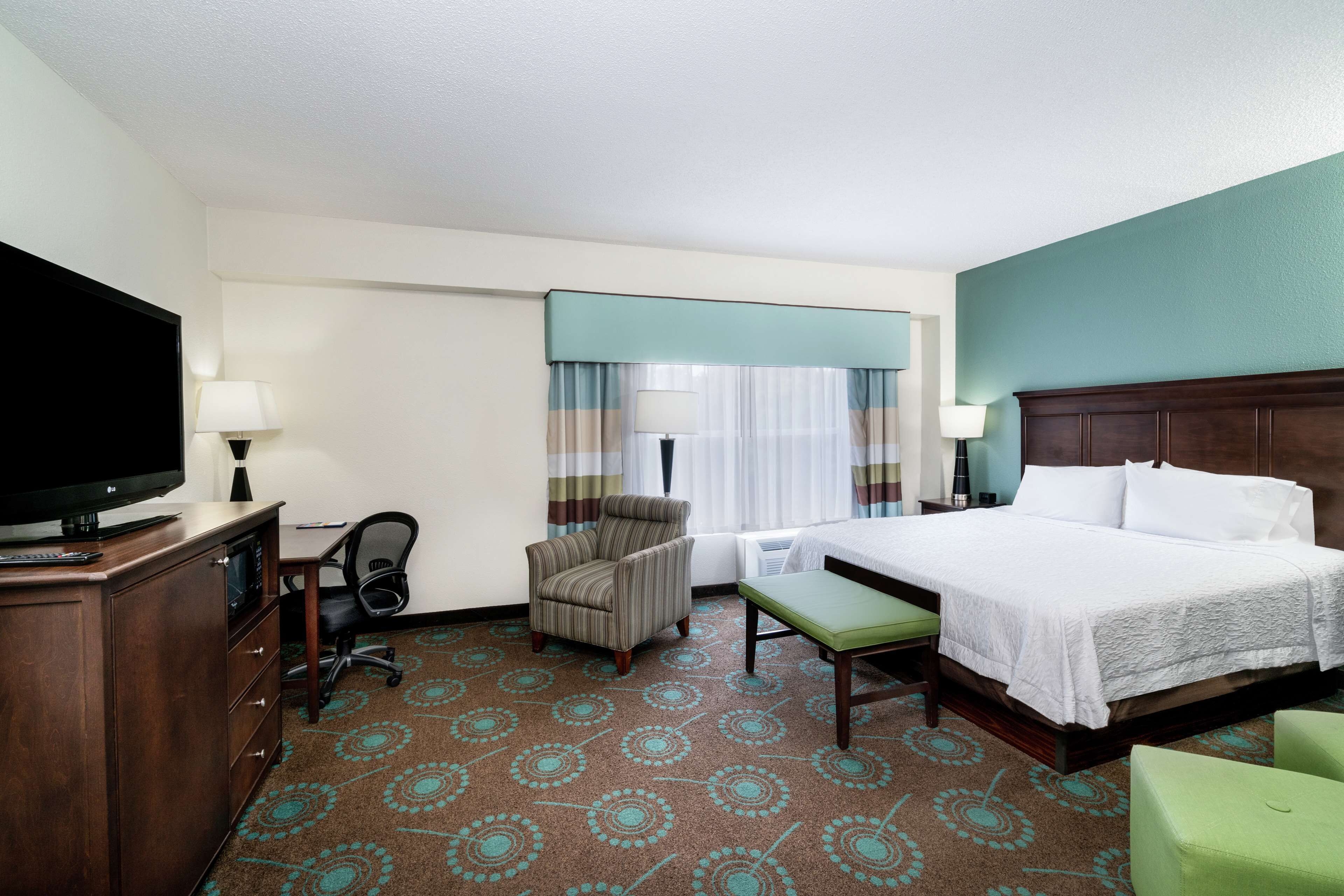 Habitación de hotel con cama, escritorio, silla, televisor y ventana con cortinas.