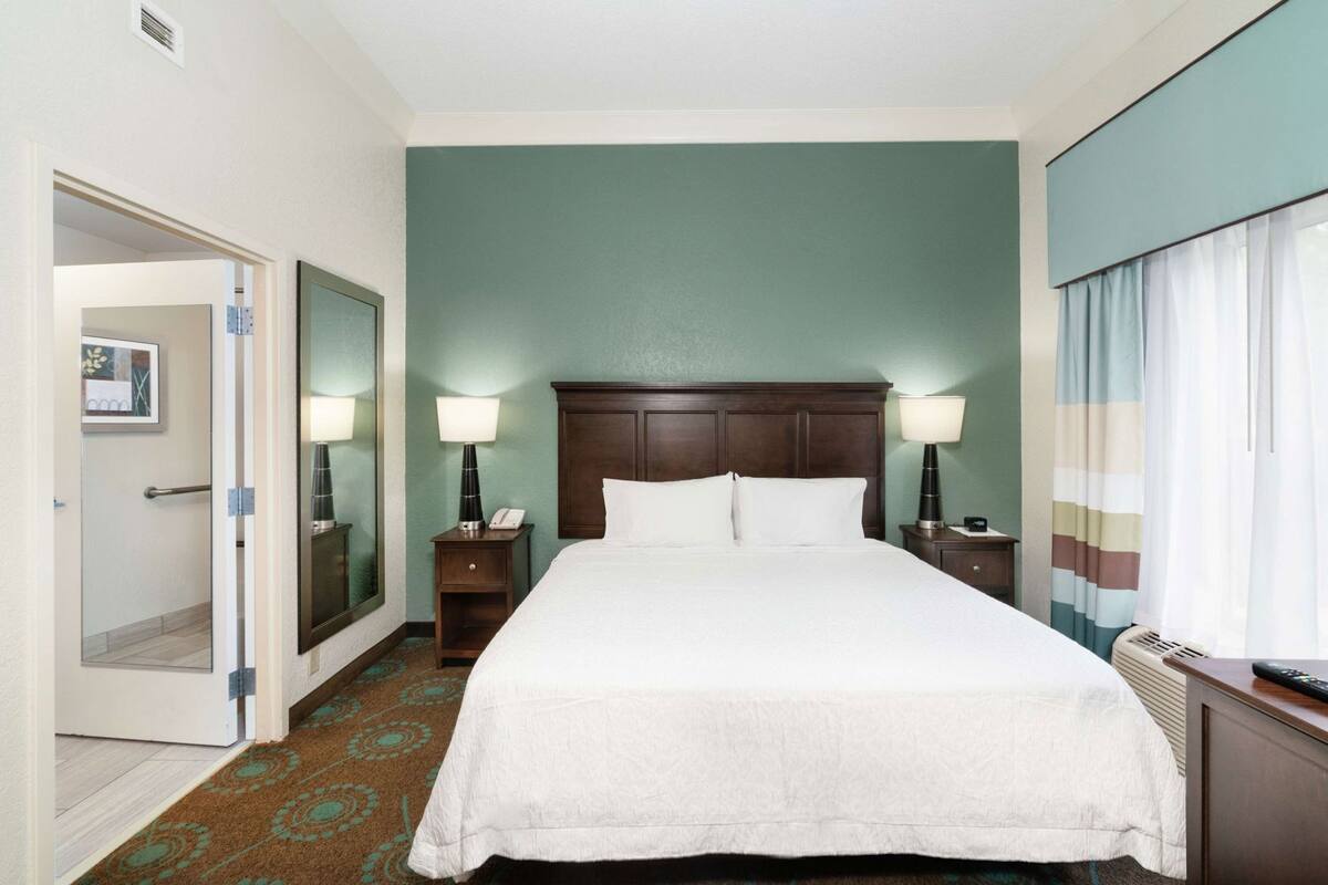 A hotel room with a large bed, bedside lamps, a nightstand, a mirror, and a view of the outdoors.