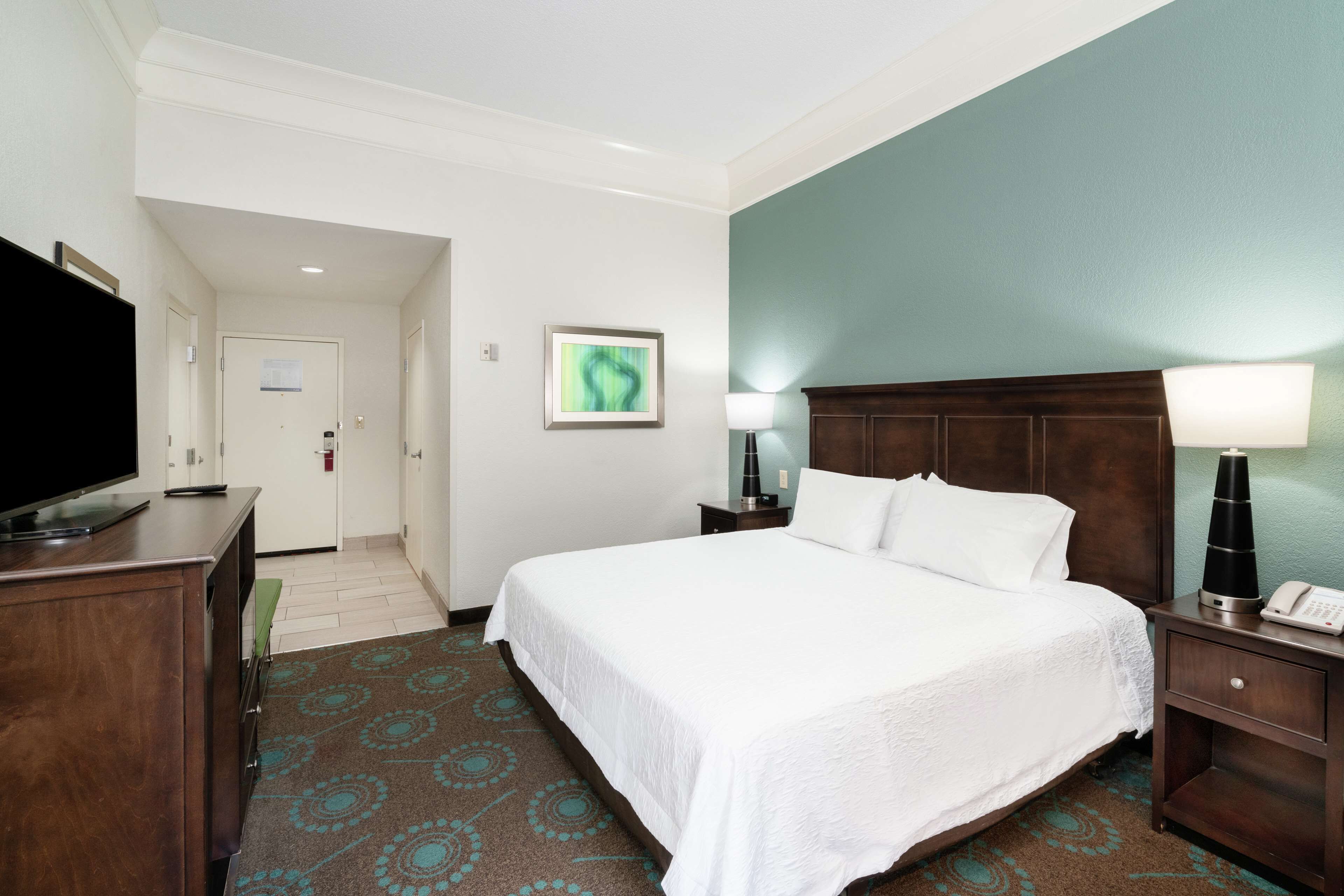 A hotel room with a large bed, a television on a wooden dresser, a nightstand with a lamp, and a framed picture on the wall.