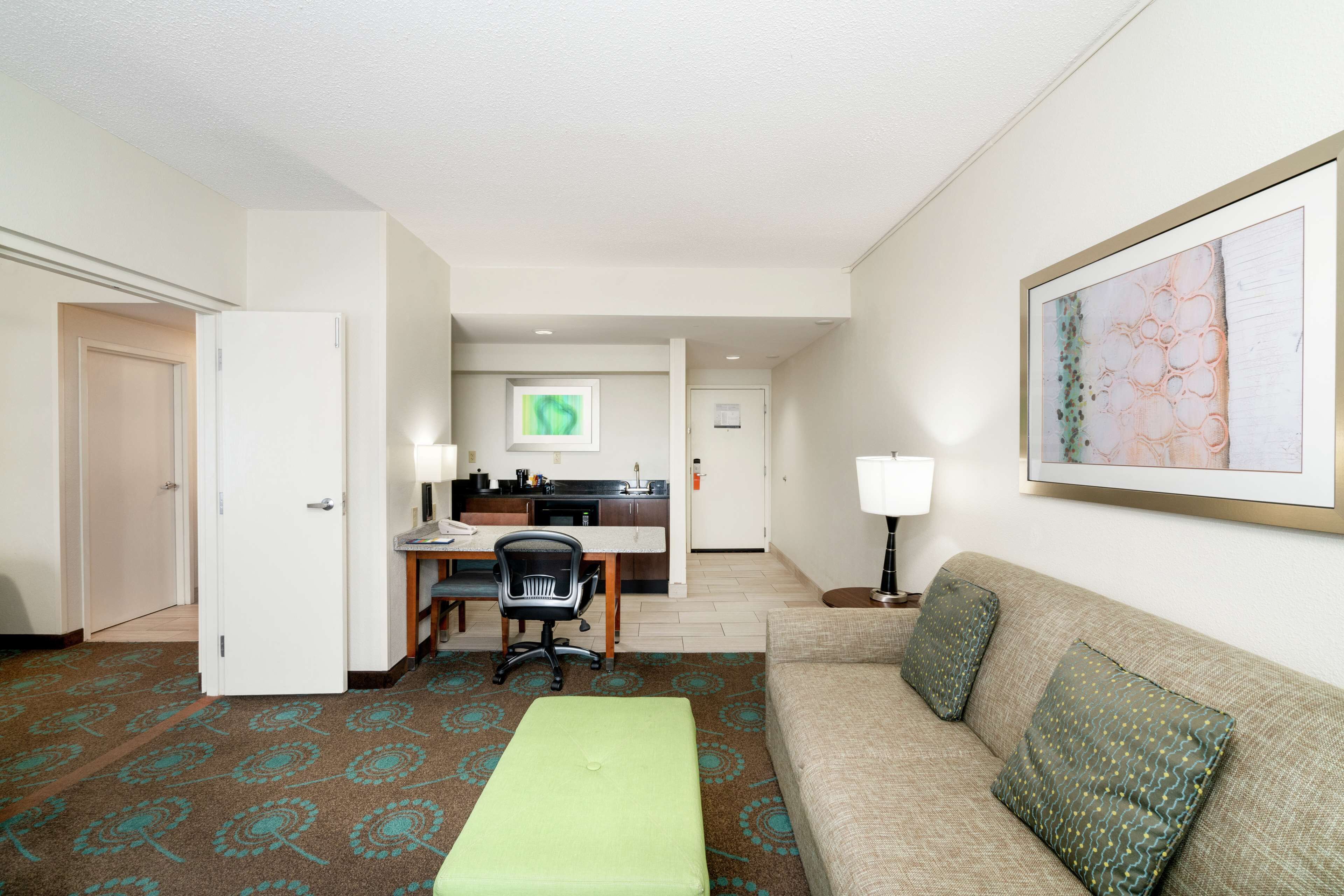 A hotel room with a green ottoman, a beige sofa with patterned cushions, a desk with a chair, a kitchenette, and a framed abstract painting on the wall.