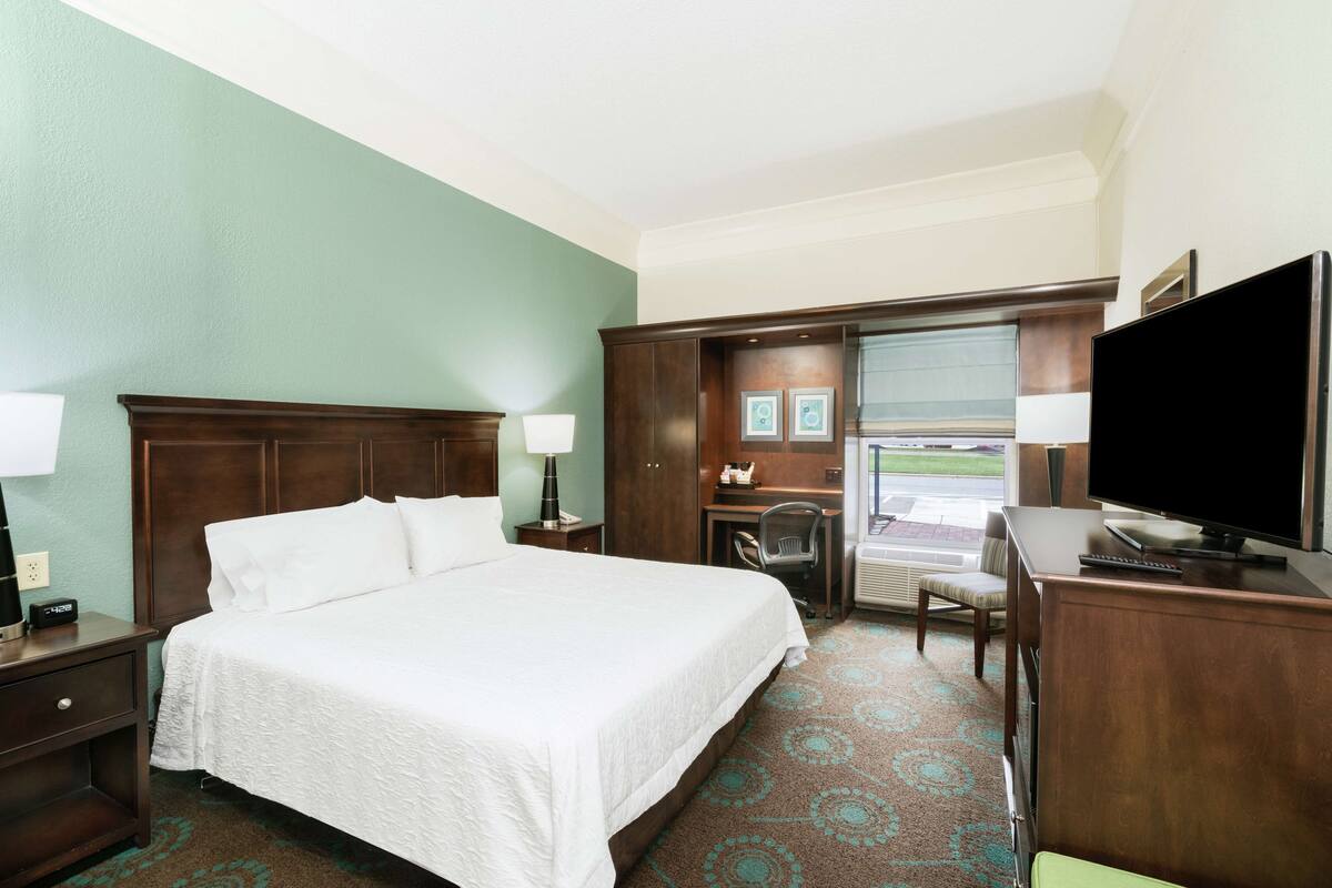 A hotel room with a large bed, a desk with a chair, a television, and a view of the outside.
