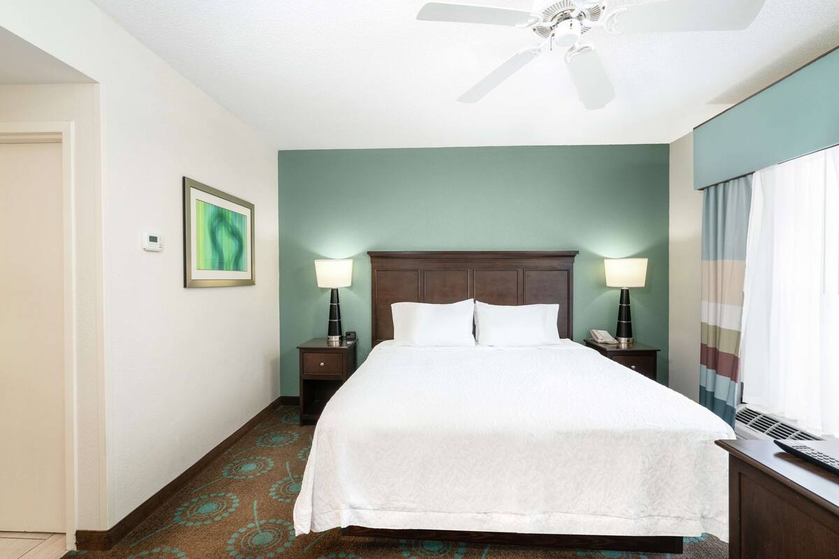 A hotel room with a large bed, two bedside tables with lamps, a ceiling fan, and a window with curtains.