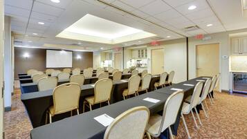 A conference room with multiple tables and chairs, a projector screen, and an exit sign.