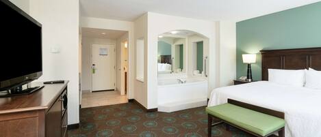 A hotel room with a bed, a bench, a TV, a desk, and a bathroom with a sink and mirror.