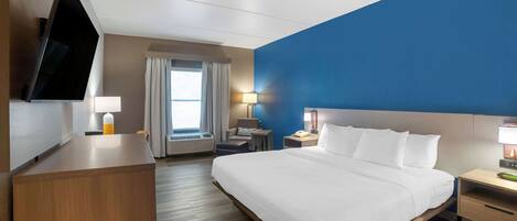 A hotel room with a large bed, a desk, a flat-screen TV mounted on the wall, a window with curtains, and a blue accent wall.