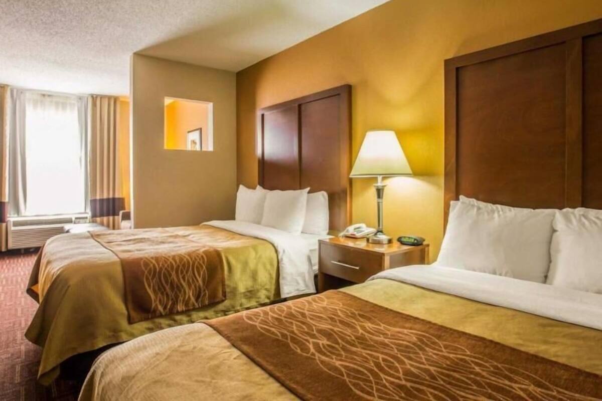 A hotel room with two beds, a bedside table with a lamp, a mirror, and a window with curtains.
