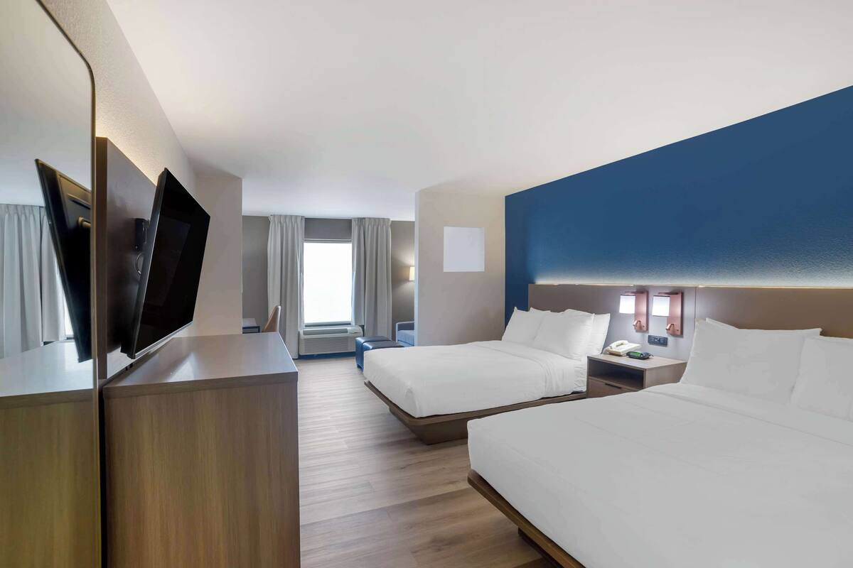 A hotel room with two beds, a desk, a flat-screen TV, a bedside table with a lamp, and a window with curtains.