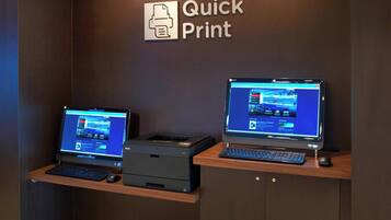 Two desktop computers on a wooden desk with a printer, a sign reading "Quick Print" on the wall, and a keyboard and mouse on the desk.