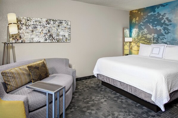 A hotel room with a bed, a gray sofa with patterned cushions, a small table, a floor lamp, and a wall art piece.