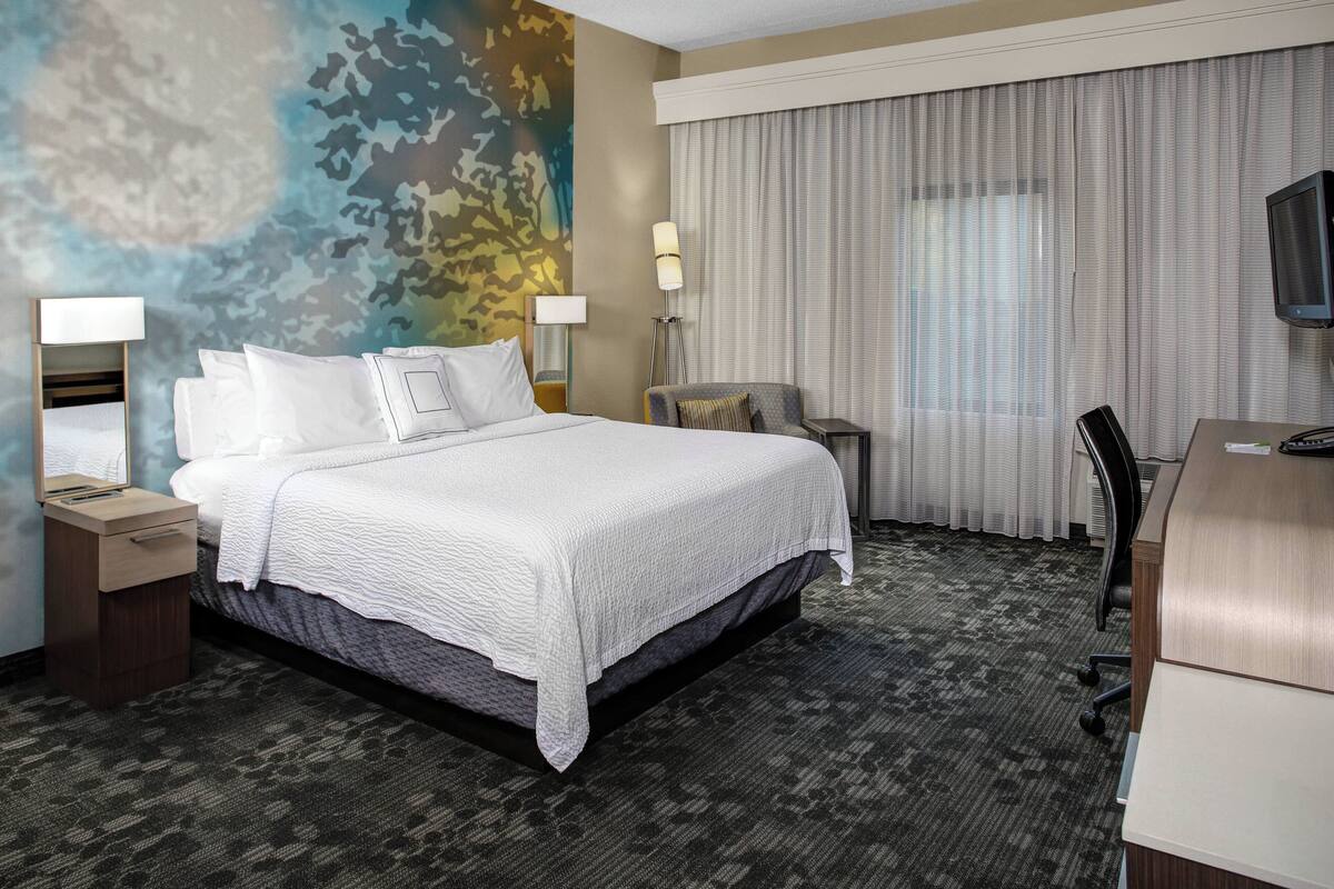 A hotel room with a large bed, a desk, a chair, two lamps, and a television.
