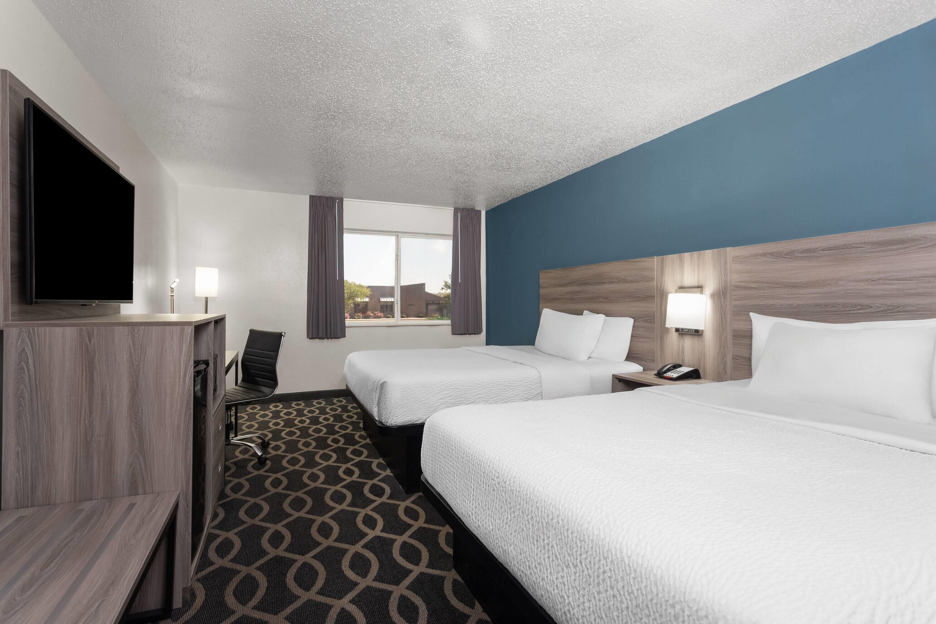 A hotel room with two beds, a desk, a flat-screen TV, and a window with curtains.
