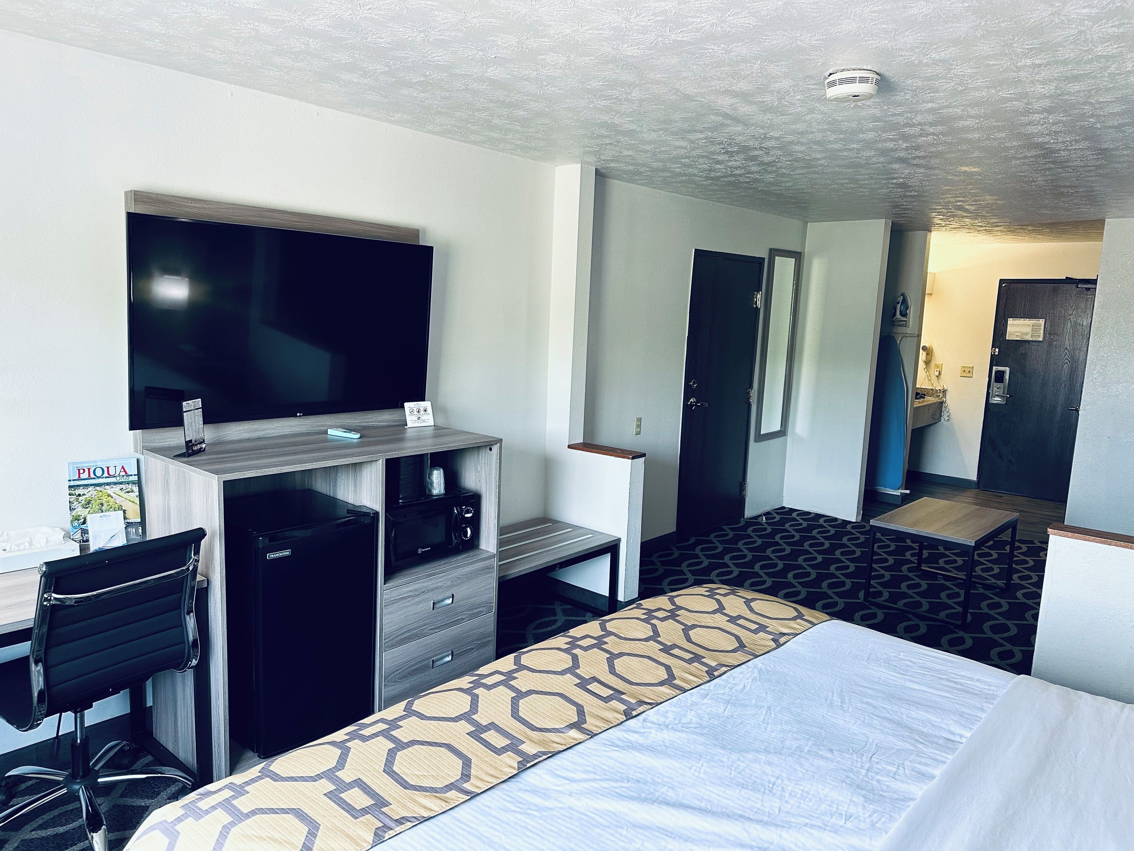 Et hotelværelse med en seng, et skrivebord med stol, et fjernsyn, en minibar og et badeværelse, der kan ses gennem en åben dør.