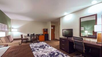 Habitación de hotel con cama, escritorio, televisor, silla, kitchenette pequeña y espejo.
