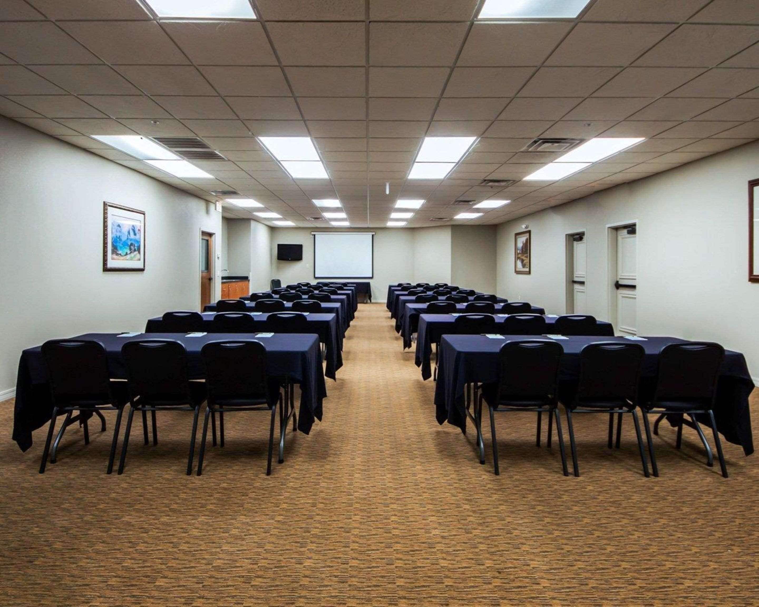 Un salón de conferencias con filas de mesas y sillas, una pantalla de proyector y cuadros en las paredes.
