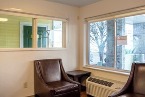 A room with two leather chairs, a small table, a window with a view of a building and trees, and a wall-mounted air conditioning unit. - HomeTowne Studios by Red Roof Salem, OR (Salem)