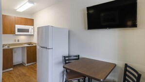 Cocina compacta con microondas, refrigerador y armarios de madera. Un televisor de pantalla plana está montado en la pared sobre una mesa de comedor con sillas.