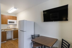 A compact kitchen with a microwave, refrigerator, and wooden cabinets. A flat-screen TV is mounted on the wall above a dining table with chairs.