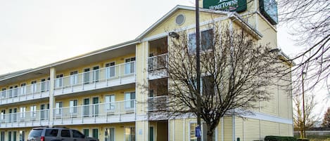 A multi-story motel with a green sign, a parking lot, and a tree in front.