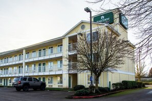 Un motel à plusieurs étages, avec une enseigne verte, un parking et un arbre devant.