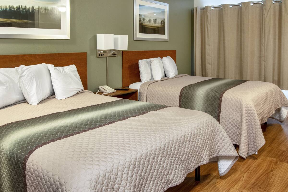 A hotel room with two beds, a nightstand with a phone, and a wall-mounted lamp.