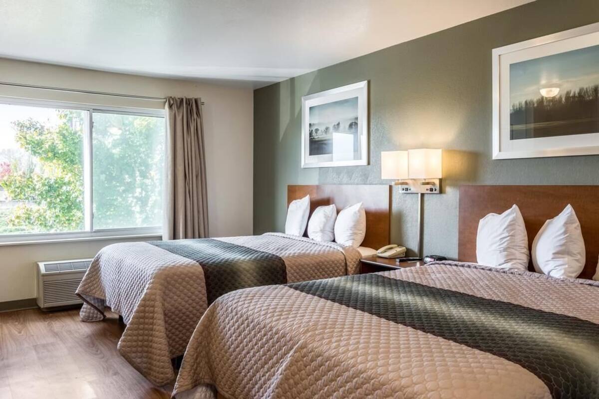 Une chambre d’hôtel avec deux lits, une fenêtre avec des rideaux, une lampe fixée au mur et des tableaux encadrés.