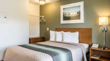 A hotel room with a bed, a nightstand with a phone, a lamp, a framed picture on the wall, and a coat rack.