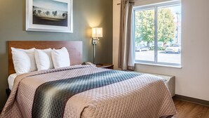 Habitación de hotel con una cama grande, una mesita de noche, una lámpara, una ventana con cortinas y un cuadro en la pared.
