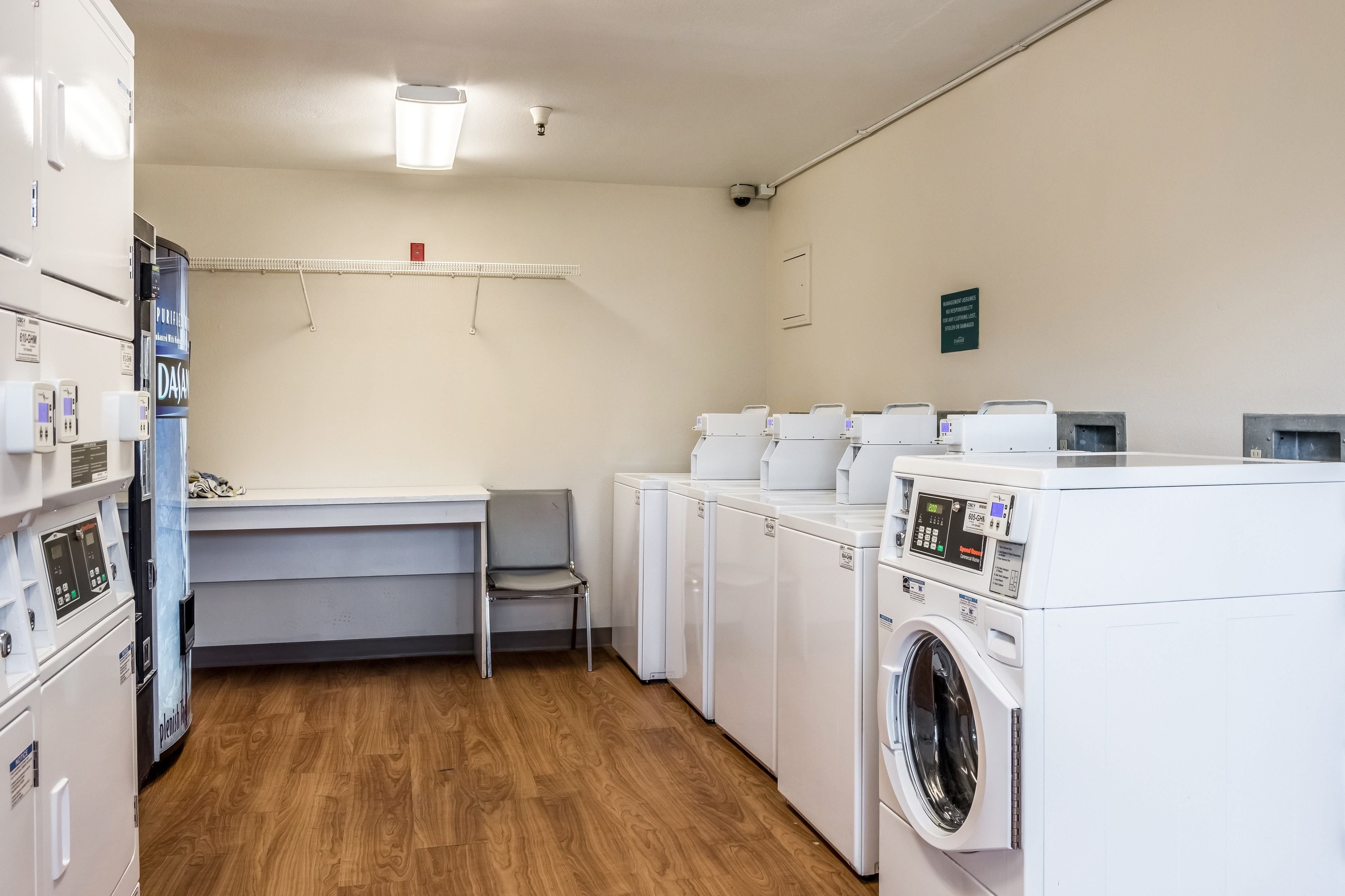 Een wasserette met een wasmachine, droger en gestapelde drogers. Er staat een stoel en een plank met een rood voorwerp erop.