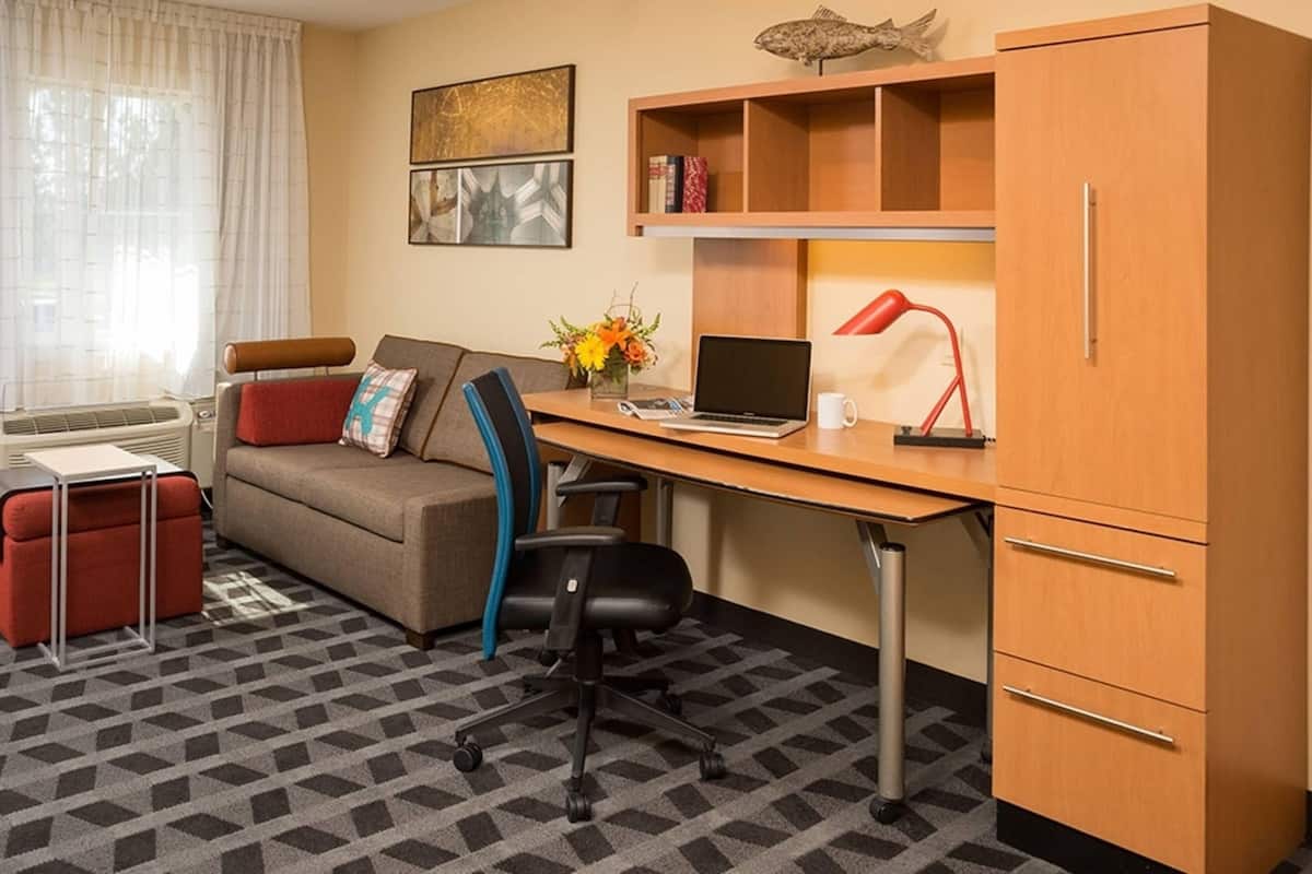 Une chambre d’hôtel compacte comprenant un canapé, un bureau avec un ordinateur portable, une lampe rouge et une petite bibliothèque.