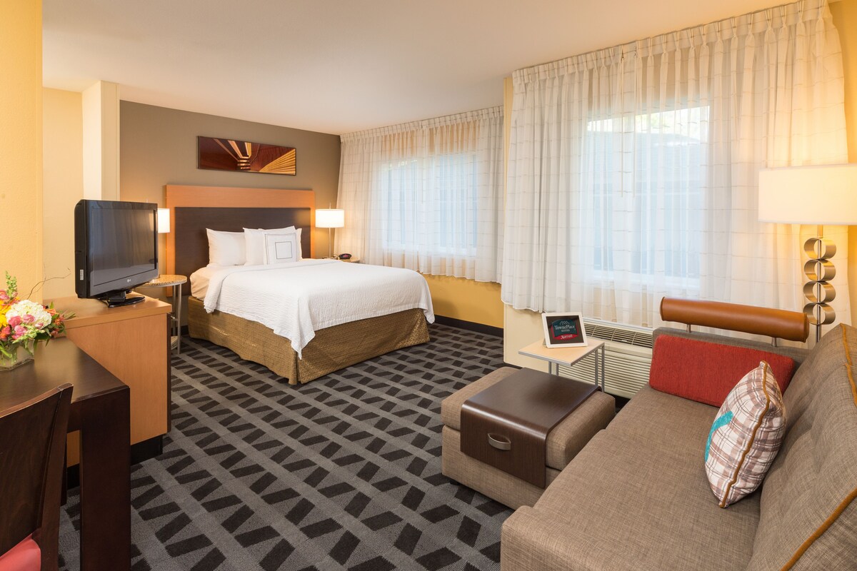 Quarto de hotel com cama, escrivaninha com televisão, sofá e pequena mesa com abajur.