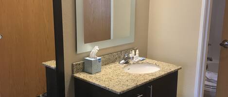 A bathroom with a wooden door, a large mirror, a granite countertop sink, and a dark vanity with drawers.