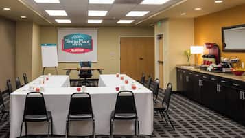 Una sala conferenze con un lungo tavolo, sedie, uno schermo per proiettore che mostra il logo di TownePlace Suites Marriott, una lavagna e un angolo cottura.