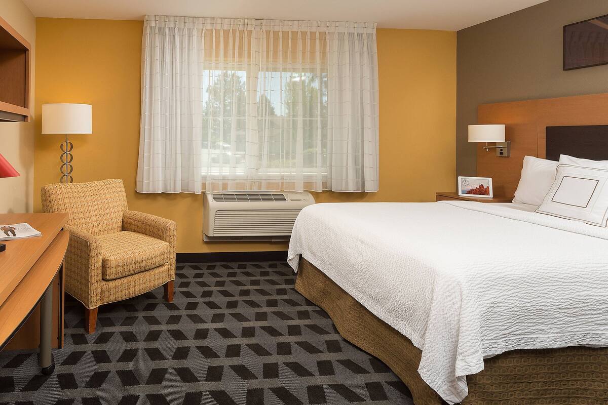 Une chambre d’hôtel avec un lit, une chaise, un bureau, une lampe et une fenêtre avec des rideaux.