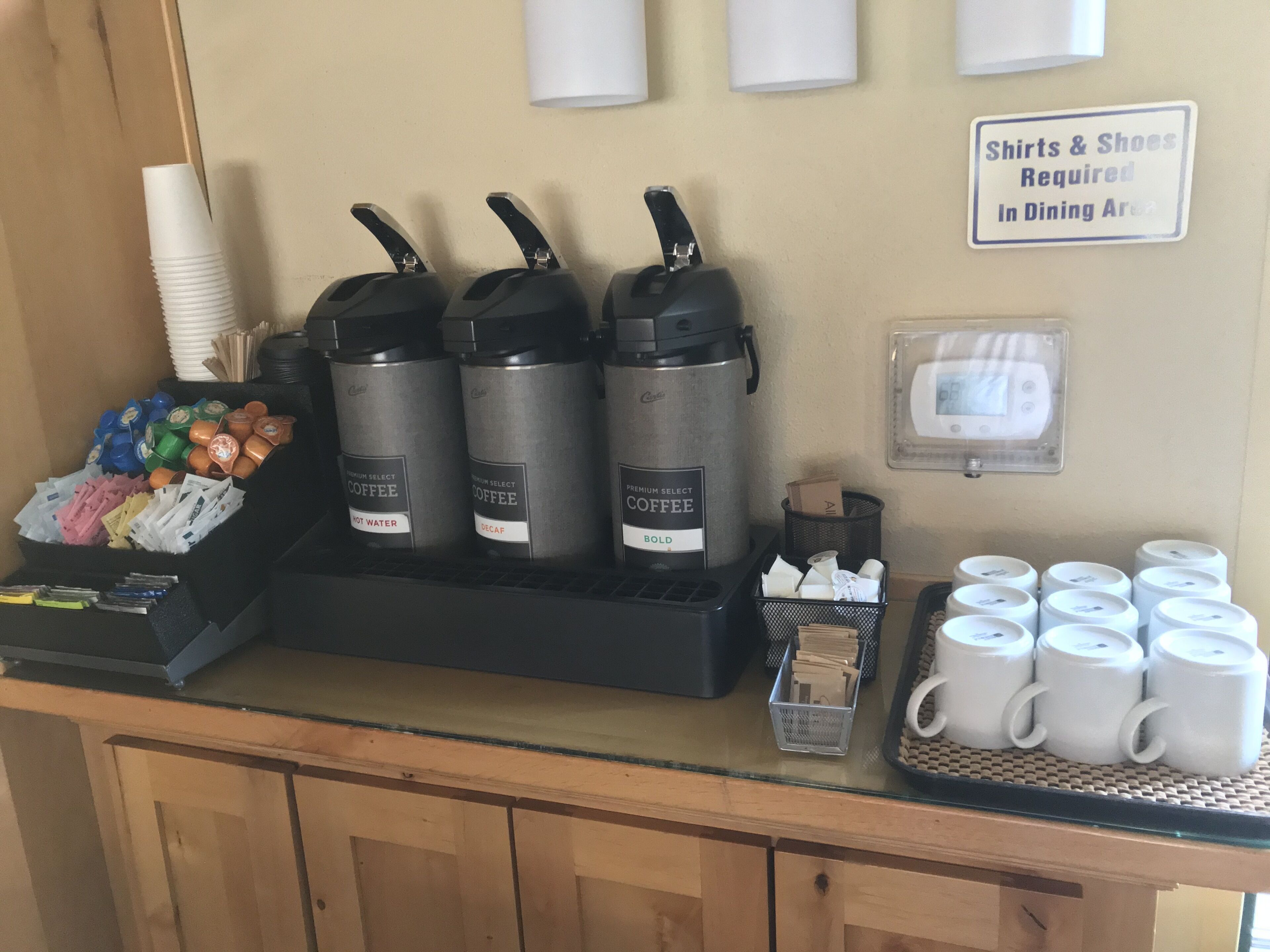 Un angolo caffè con tre macchine da caffè, un vassoio di bicchieri usa e getta e un cartello che indica l'obbligo di indossare camicie e scarpe nella sala da pranzo.
