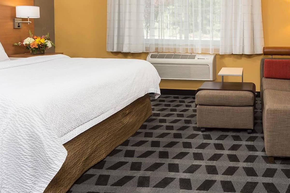 Une chambre d’hôtel avec un grand lit, un petit canapé, un appareil de climatisation, une table de chevet avec une lampe et une fenêtre avec des rideaux légers.